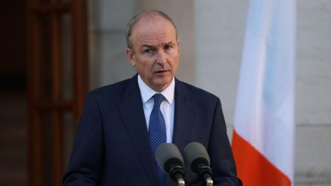 Micheál Martin is wearing a navy suit and tie with a white collared shirt. He stands at green podium which has two black microphones attached to it. Behind his left shoulder is an Irish tricolour. Behind his right shoulder is a brown door to a cream-brick building.