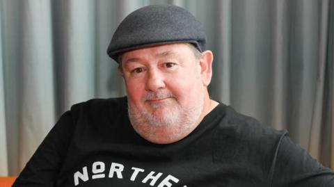 Johnny Vegas wearing a dark flat cap and a black T-shirt with the word “NORTHERN” printed in bold white letters across the chest. The background consists of grey curtains.
