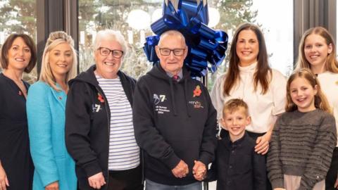 Eight people, some of them children, pose for a photo in front of some glass doors and a large blue ribbon.