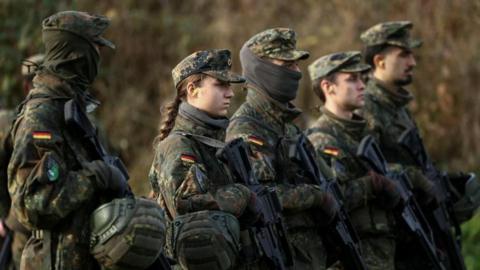 Male and female soldiers stand with guns in a field, wearing camouflage with the German flag stitched on their upper arms.