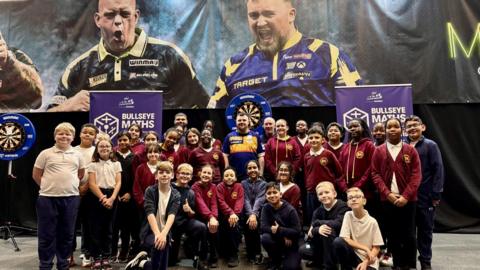A group of school children standing infront of darts boards and some adults are also in the photo. They are smiling and some have their thumbs up. Their school uniform is maroon with a yellow crest.