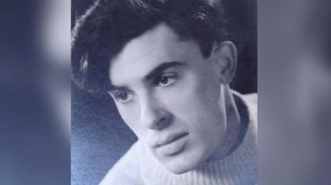 Peter Gibbs as a young man in a black and white image. He has thick black hair and is wearing a white fisherman's sweater.