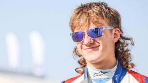 George has a brown mullet and is wearing blue tinted sun glasses whilst in his overalls.