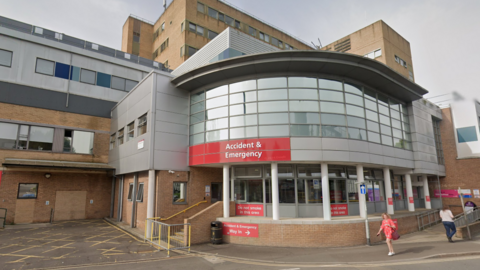 The outside of Yeovil District Hospital's Accident & Emergency department. The reception is a curved, grey cladded building, with some orange brick sections, and red signs. The larger portion of the hospital is seen behind.