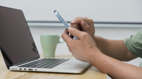 A man using a mobile phone in front of a grey laptop placed open on a table - we can only see his arms. A green mug is in the background.