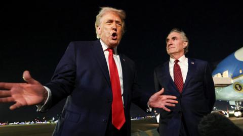 US President Donald Trump on the airport tarmac with Air Force One behind him. The president has his hands and mouth open as he speaks to a boom mic. Secretary of the Interior Doug Burgum stands to the president's left and watches him speak.
