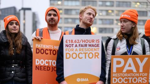 Resident doctors wearing orange beanie hats and carrying orange placards bearing the wears Doctor Need Jobs