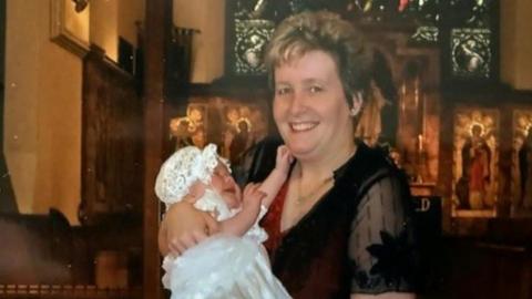 A scanned image of a middle-aged woman with short hair. She is standing in a church wearing a red dress with a mesh black top over the top. She is holding a crying baby wearing a white Christening dress and hat.