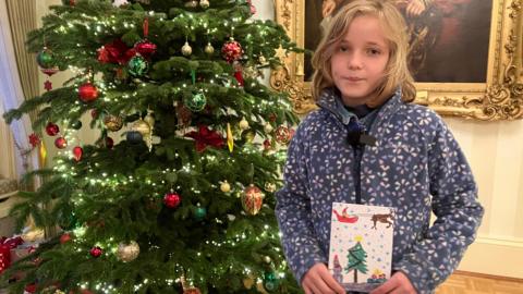 A child holding a Christmas card she has drawn standing in front of a Christmas tree in a Government building.
