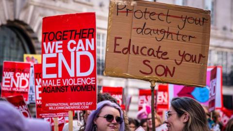 Women at a protest in London to mark International Women's Day hold placards reading "together we can end male violence against women" and "protect your daughter, educate your son".