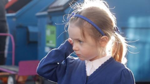 Four-year-old Beatrix in her blue school uniform, stood outside school in the morning sunshine on her first day.
