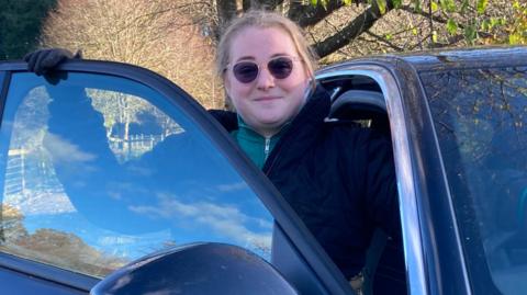 Dynes yn gwisgo siaced du a sbectol haul yn dringo allan o car mewn lleoliad gwledig