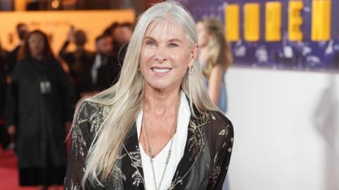 Sharron Davies wearing a sheer black jacket with metallic floral patterns over a white blouse, paired with light-colored trousers. The individual is standing on a red carpet at an event, holding a light handbag.