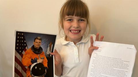 A young girl smiling, holding a picture of an astronaut in one hand and a letter in the other. She wears a white T-shirt
