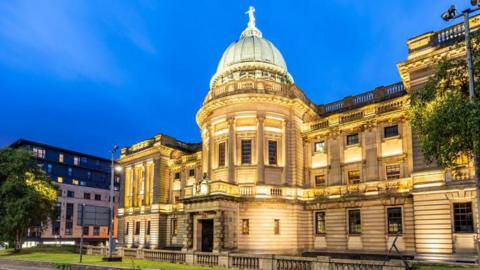 Large lit up library on a street at night time. Library has a dome style light blue roof and the library itself is a sandstone yellow lit up by lights