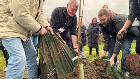 Three men are planting the sapling in muddy ground. To the left, one man is holding up dark green tarpaulin to shake off soil into the hole where the tree sits. Only his body and legs are visible. Two other men - one older, with white grey hair, beard and glasses - and a younger man with dark blonde hair tied back in a ponytail - are also hauling up the tarpaulin to tip the soil out. The thin sapling is attached to a tube of pale green protective plastic. A brown dog peers into shot from the right.