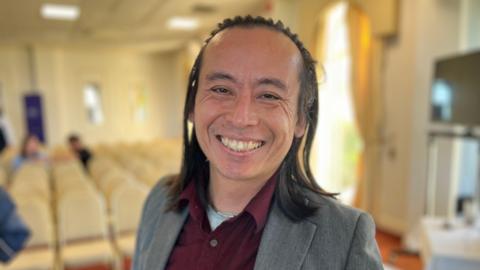 Deputy David Goy smiling at the camera. He is wearing a grey suit jacket and a burgundy shirt. In the background are rows of white seats. He has dark shoulder-length hair.
