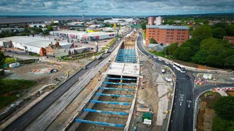 Aerial view of a roadworks site. The road is completely dug up in the middle of the carriageway with blue steel supports attached to the sides of the road as they dig down to a lower level.