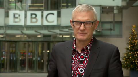 Jeremy Vine stands outside a BBC building, he wears a red and white patterned shirt under a black suit jacket. He has short grey hair and wears black rimmed glasses