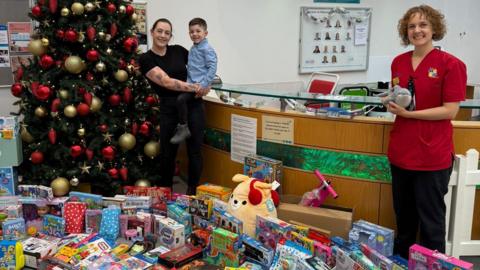 Polly, a woman, wearing a black shirt and black trousers, holding her son in her arms, standing in front of a Christmas tree with boxes of presents in front of them and a member of the children's ward, a lady in red shirt smiling for camera with them.