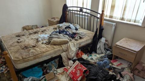 An unmade bed with a mattress covered in animal faeces and large amounts of household waste covering the floor