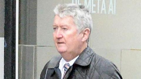 Andrew Hyams, a man with short grey hair wearing a white shirt, striped tie and black padded jacket, walks past a building.