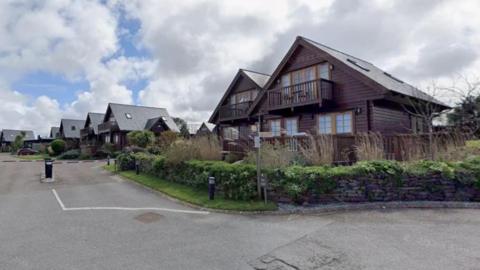 The image shows several wooden holiday lodges with pitched roofs. There are seats on the verandas and a balconies on the first floor of the lodges, which are surrounded by shrubs.