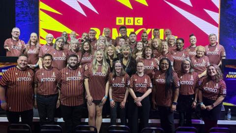 A large group of people is standing together in front of a brightly lit stage backdrop featuring the BBC logo and bold red, yellow, and white graphic patterns. The individuals are wearing matching striped shirts with the word “JCT600” printed on them.