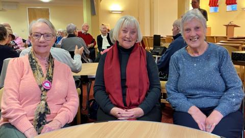 Christine wears glasses and has short grey hair and sits on the left wearing a pink top with a scarf around her neck. Rita sits in the middle with grey hair almost down to her shoulders and wears a thick red scarf and black top. Jenny sits on the right wearing a sparkling blue top with short grey hair.