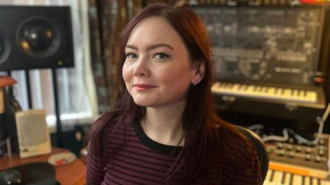 Angie smiling looking at the camera. She has brown hair and is wearing a striped black and burgundy top. In the background, there are two keyboards and a speaker.