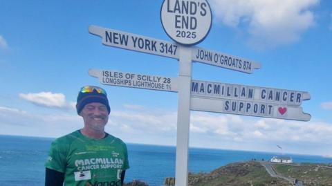 Ben Dobson pictured at Lands End, Cornwall, during his running challenge. Ben is wearing a green Macmillan Cancer Support t-shirt, a hat, sunglasses, black shorts and black running leggings.