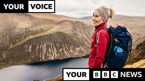 Woman with a rucksack on her back looking over a valley in the Highlands of Scotland. She is wearing a red fleece and has her hair tied back. There are rugged hills in the background and a loch at the bottom of the valley.