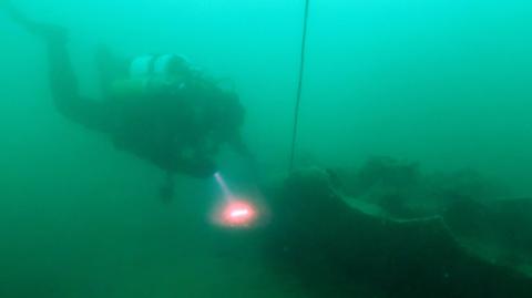A diver with air tanks on their back shines a light, red with a white glow at the centre, as they near a long rocky looking base, the remains of a shipwreck. They are in green water, what appears to be a cable extends upwards from the wreck.