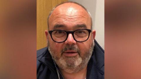 A close up photo of taxi driver Geoff Scott. He has a short, greying beard and wears black-rimmed glasses and a navy jacket.