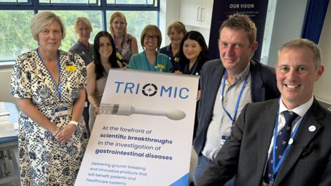 Seven women and two men are standing in a group, smiling at the camera. They are inside a hospital, and between them is a large white posted board with the word "Triomic" on the top. There is a picture of a white medical device on it that collects rectal mucus samples.