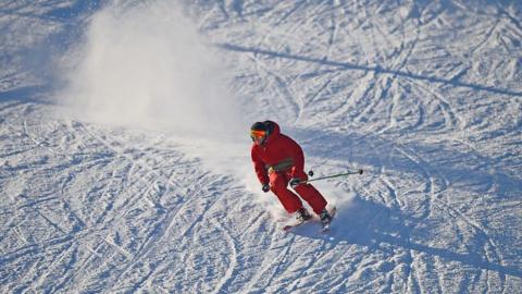 A skier dressed in red skiing gear going down a snowy slope. He is wearing a hat and ski goggles and is carrying poles. There is a cloud of snow behind him.