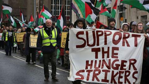A large crowd of protesters carrying Palestinian flags with a row at the front holding a large white banner which reads 'support the hunger strikers' in red lettering.