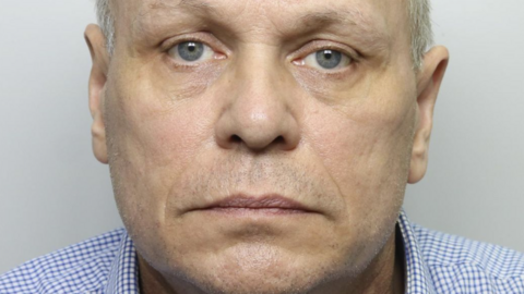 Police custody image of a man in his late 50s. He has a neutral expression and looking at the camera. He is wearing a blue shirt.