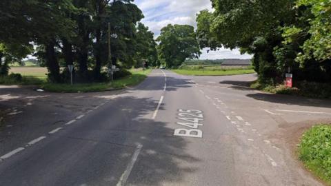 Google maps image of a crossroads with three different turnings. There is green grass and trees beside the road.