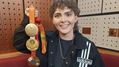 A woman with curly brown hair holds up three gold medals. She is wearing a a black t-shirt, a black Adidas jacket, a gold chain around her neck.