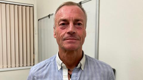 Andrew Tabel looking directly into the camera. He's wearing a blue checkered shirt and in the background there is a whiteboard.