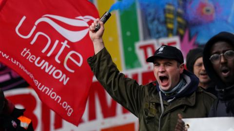 A striking bin worker wears a black cap and green coat. He is shouting and holding a blue flare in his right hand. He stands next to a red and white Unite the union flag