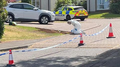 Police tape outside a suburban home with a paved driveway. A police car is parked on the corner and a forensics officers kneels examining a patch of ground.