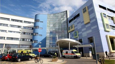 A picture of the John Radcliffe Hospital in Oxford. It is blue and grey, are there are cars and ambulances parked in front of it.