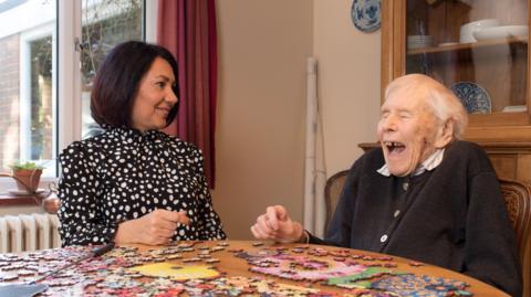 Two women - one very eldely - laugh as they make a puzzle. The old woman is missing teeth.