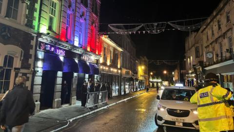 A night scene on a busy street with a police officer in the corner and people waiting outside a venue