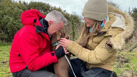 Chris Feenery (left) and Leanne Allcote are crouching in a field facing each other, holding Toffee, the brown dachshund. They are wearing outdoor coats, and Leanne is wearing a woolly hat. Chris has his head buried into Toffee's and Leane is smiling.