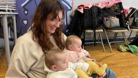 The photo shows a mum with shoulder-length, chestnut hair, smiling, wearing glasses, a cream sweatshirt and jeans, sitting down on a wooden floor with her twin baby daughters sitting on her lap. One baby is wearing a pink cable knit cardigan over a yellow babygrow, while the other is wearing a cream hooded, knitted top.