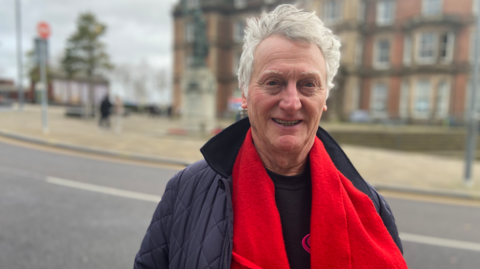 A man with grey hair in a blue jacket, wearing a red scarf, smiles at the camera. He is standing outside in front of a large building, which is on the other side of the road he is standing next to.
