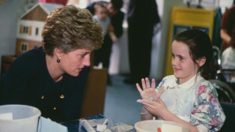 Princess Diana, wearing a blue Chanel suit, speaks to a young girl playing with play-doh. The girl, who is looking at Diana, is wearing a floral jumper and has a sphere of play-doh in her hands.
